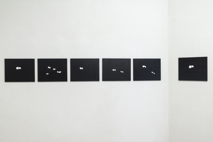 Exhibition installation: small black paintings hung in a line on a wall text on the floor