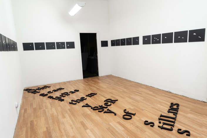 Exhibition installation: small black paintings hung in a line on a wall text on the floor