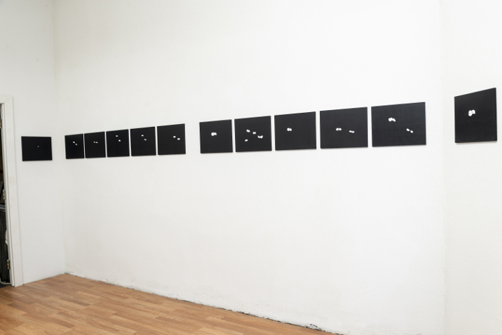 Exhibition installation: small black paintings hung in a line on a wall text on the floor