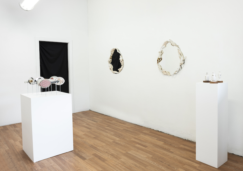 Installation shot of exhibition wall mirrors on the right handheld mirrors on a pedestal in the center. small perfume bottles on smaller pedestals