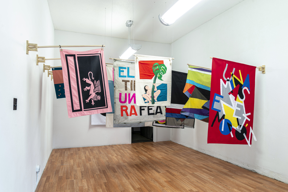 installation view: colorful flags hang in a clean well lit space with wooden floors and white walls