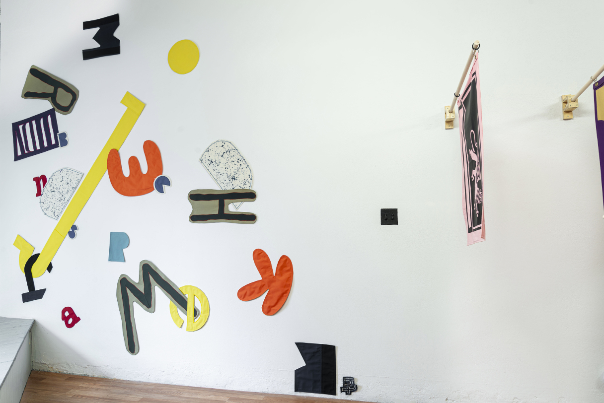 installation view: colorful flags hang in a clean well lit space with wooden floors and white walls