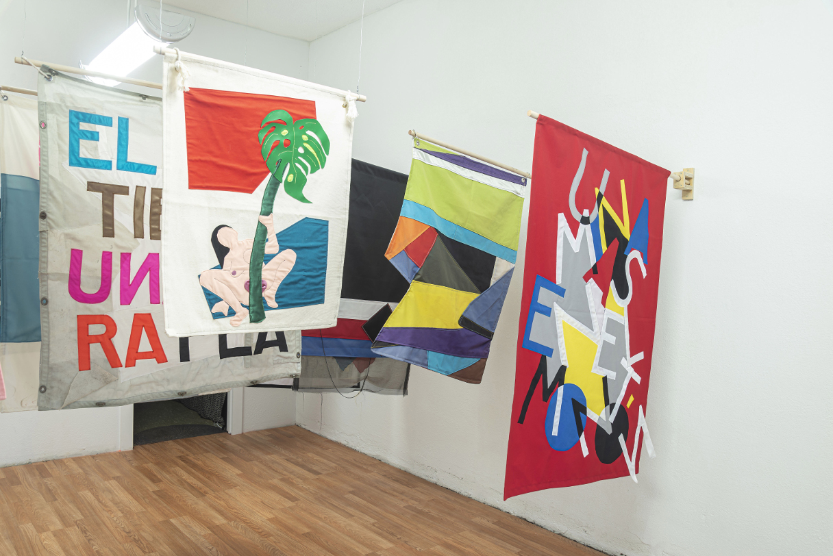 installation view: colorful flags hang in a clean well lit space with wooden floors and white walls