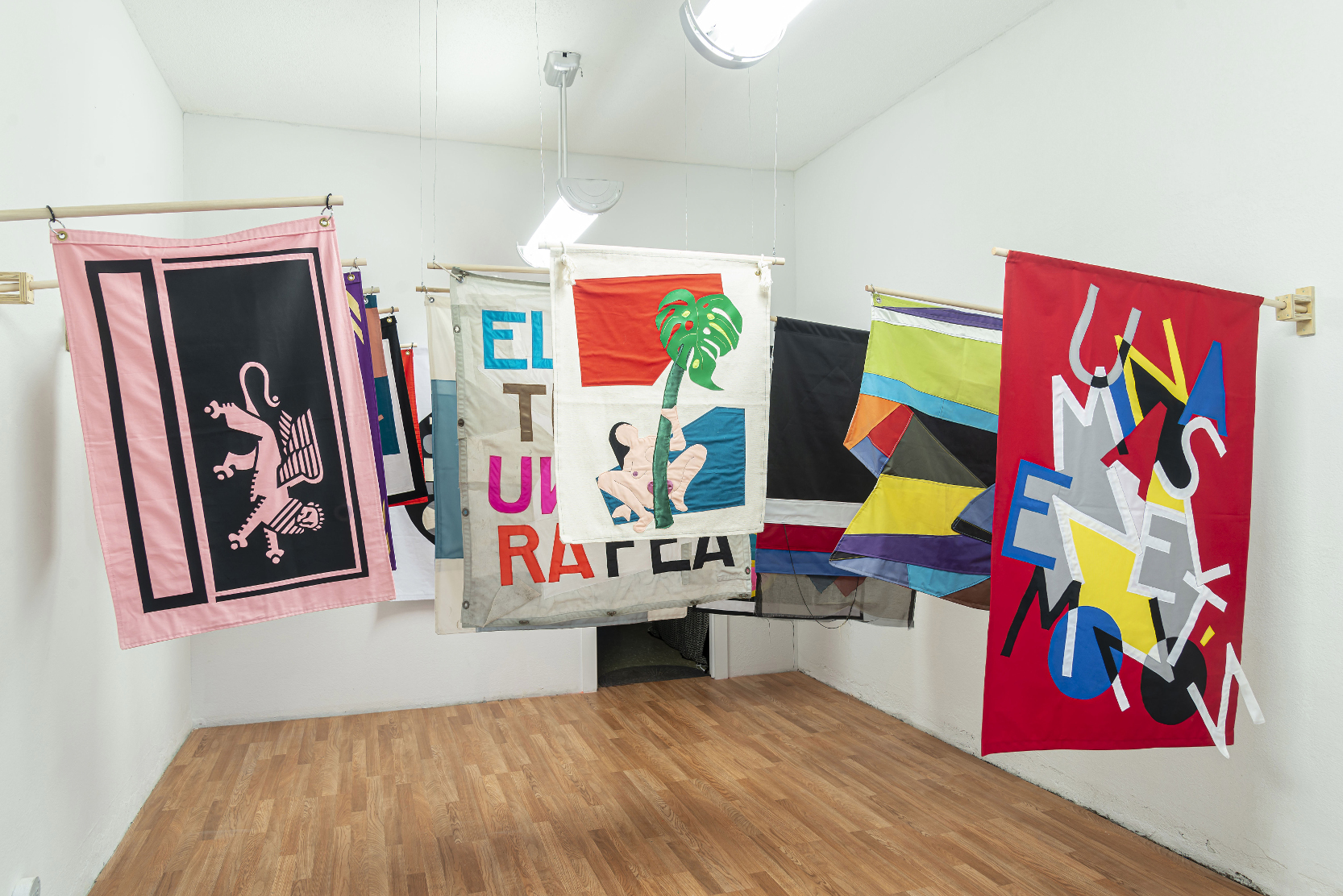 installation view: colorful flags hang in a clean well lit space with wooden floors and white walls